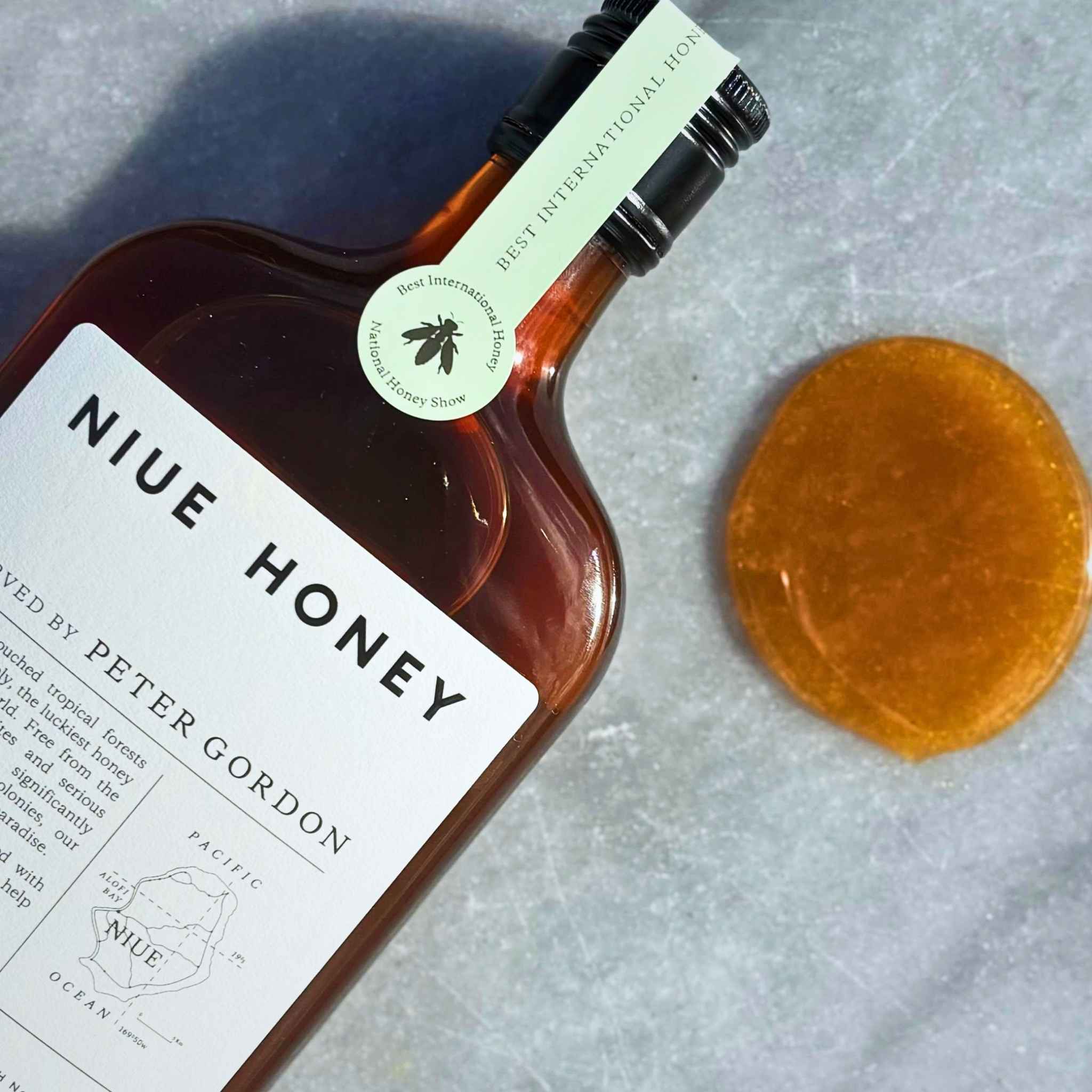 A bottle of Niue Honey by Peter Gordon with a white label and black cap, placed diagonally on a textured gray marble surface next to a round drop of the amber Niue Honey.