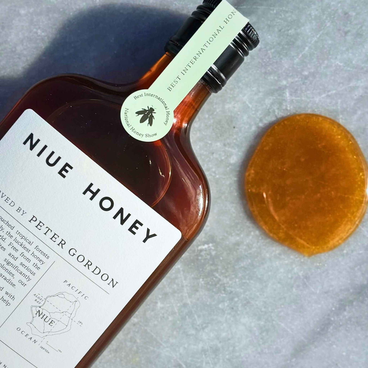 A bottle of Niue Honey by Peter Gordon with a white label and black cap, placed diagonally on a textured gray marble surface next to a round drop of the amber Niue Honey.