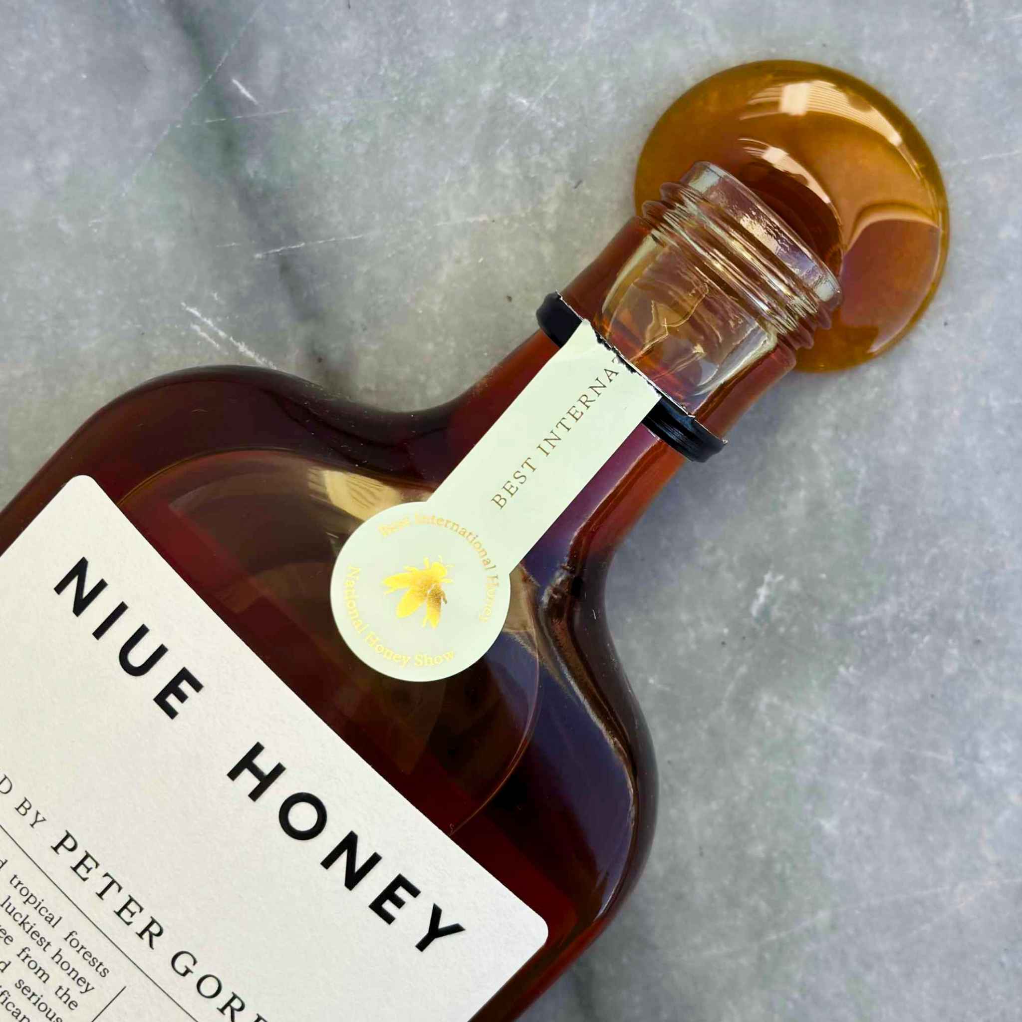 A close-up of a bottle of Niue Honey with amber liquid pooled at the neck on a light gray stone surface. The label reads 'NIUE HONEY' and 'BEST INTERNAL' with a small bee emblem.