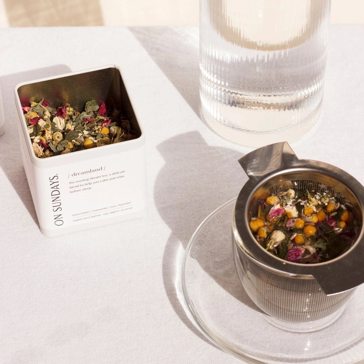 On Sundays Loose Leaf Tea Infuser in use with Dreamland organic loose leaf tea, surrounded by chamomile, rose, and botanicals, next to its white square tea tin on a sunlit table.