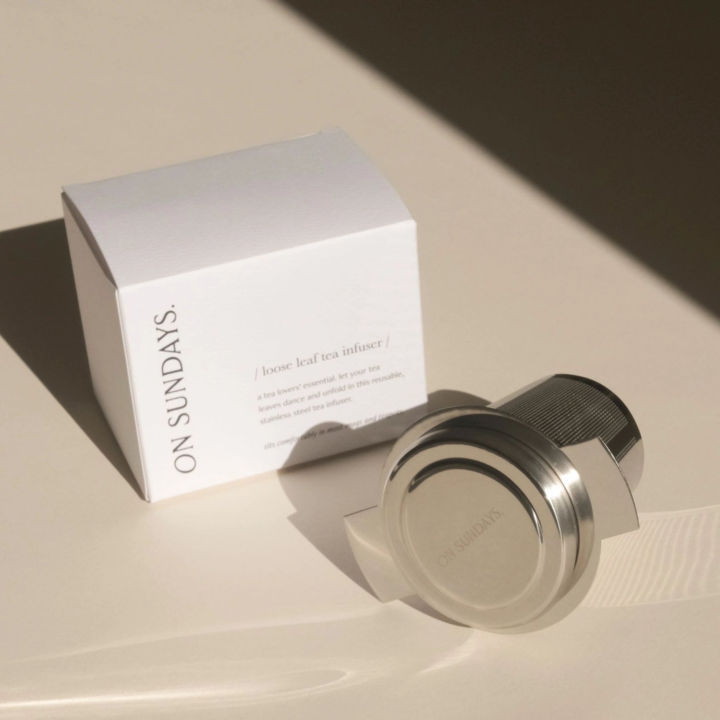 On Sundays Loose Leaf Tea Infuser with stainless steel lid shown beside its minimal white packaging, photographed in soft natural light on a neutral-toned surface.