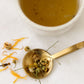 A spoonful of dried chamomile, calendula, and lavender beside a cup of brewed Unwind tea—organic botanicals crafted for a calming bedtime ritual.