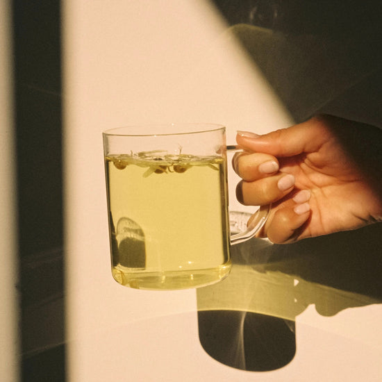 Hand holding a calming cup of On Sundays tea Unwind blend, brewed to a soft golden hue, captured in natural light for a relaxing wellness moment.