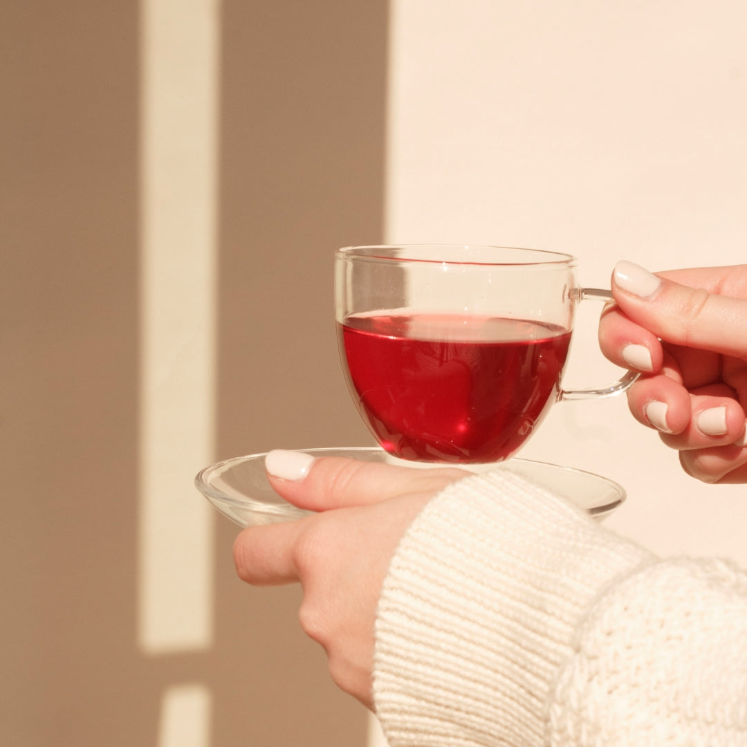 Hands holding a glass cup of On Sundays Mulled Wine organic tea with rich red herbal infusion in soft sunlight.