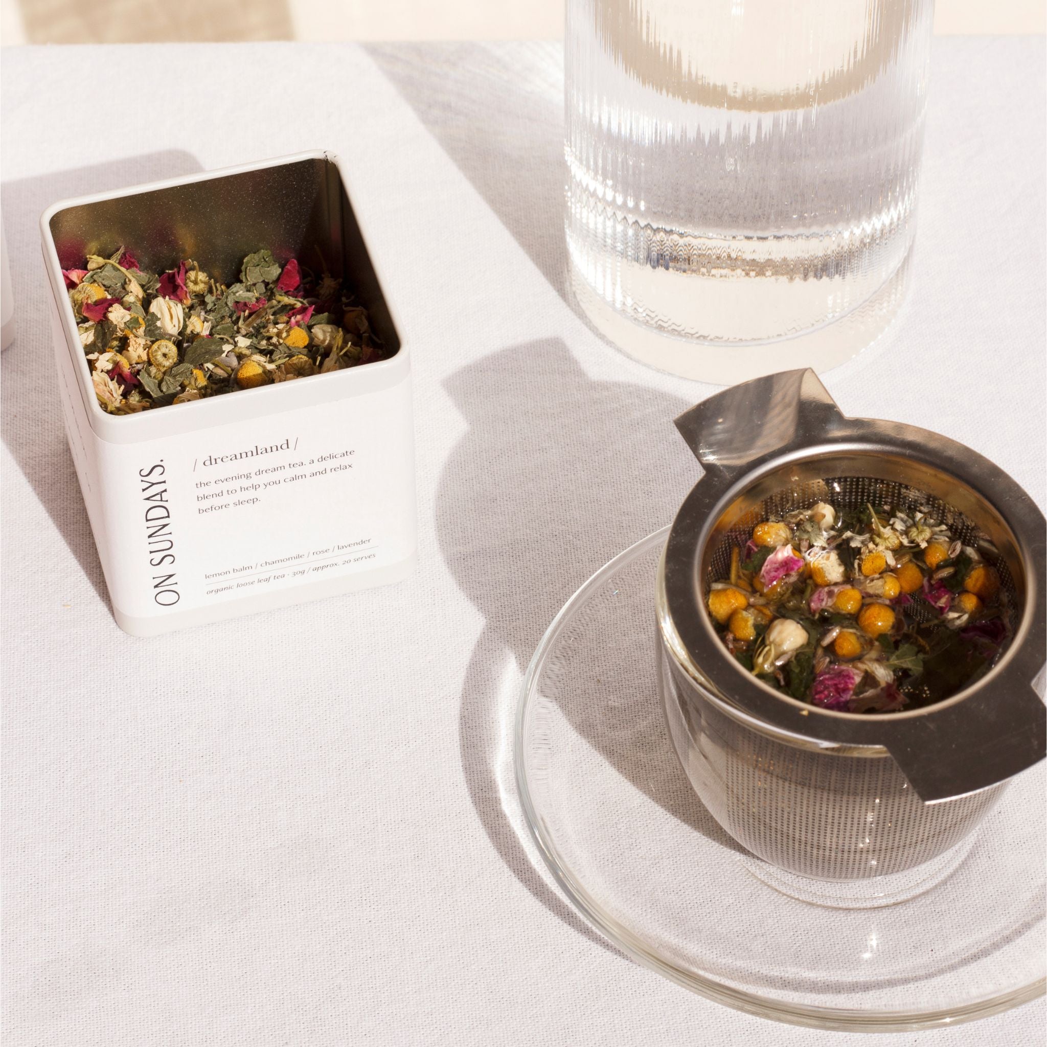 On Sundays Dreamland organic loose leaf herbal tea in white storage tin with stainless steel tea infuser brewing chamomile, rose, lavender and lemon balm blend beside glass water carafe.