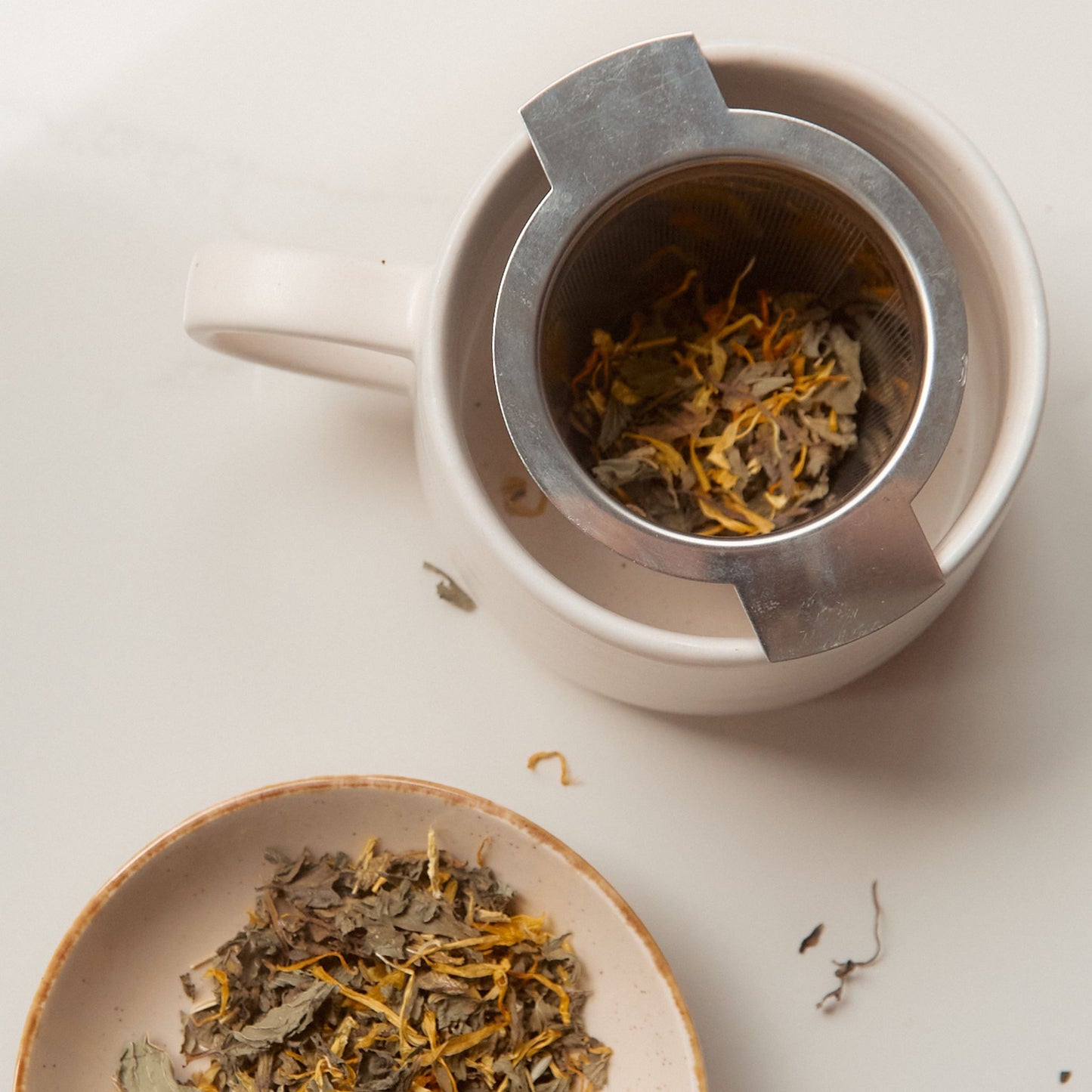 On Sundays loose leaf tea infuser filled with organic herbal tea leaves resting in a ceramic teapot with the On Sundays Ease loose leaf blend in bowl below.