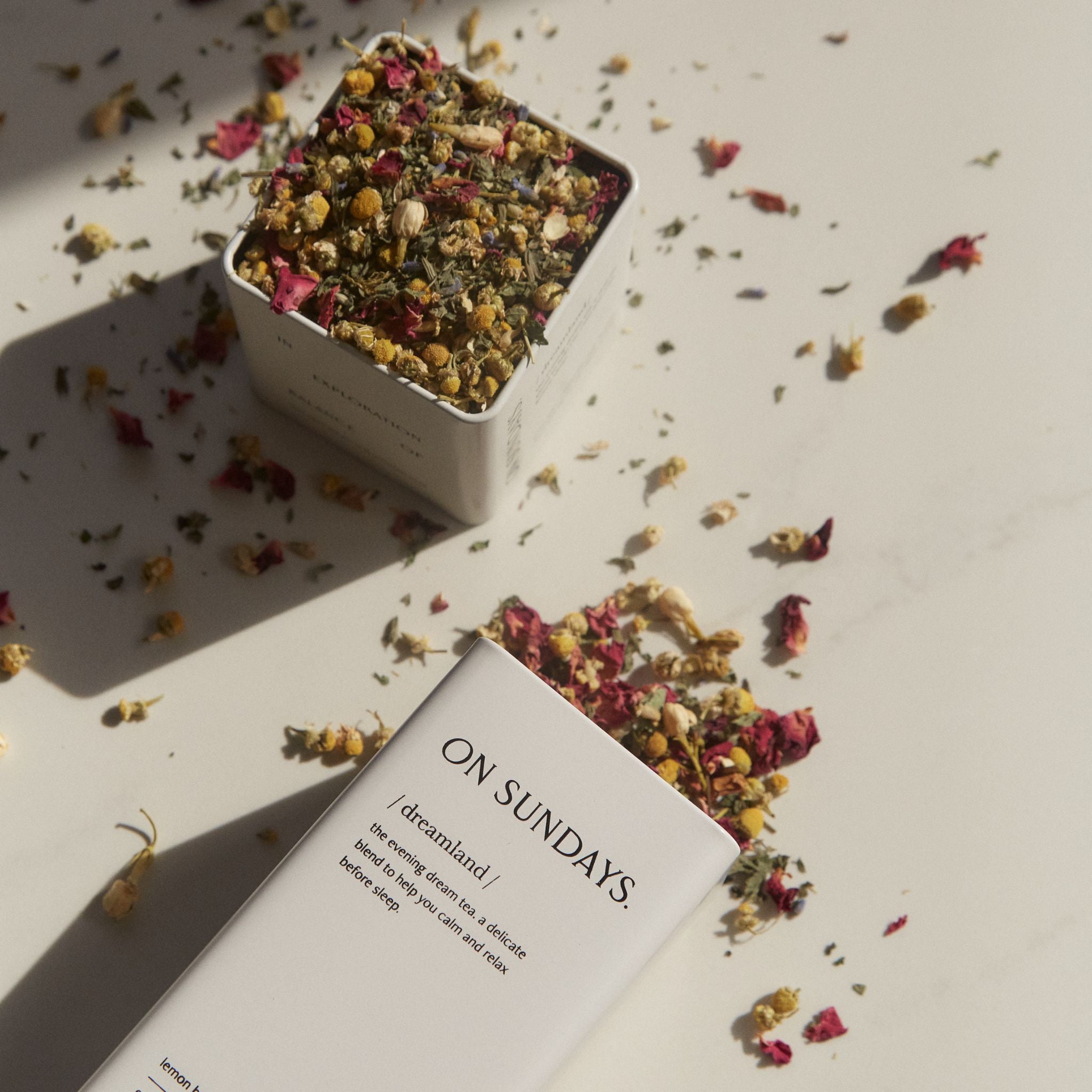 Two full tins of On Sundays Dreamland organic loose leaf sleep tea with chamomile rose and lemon balm scattered on surface.