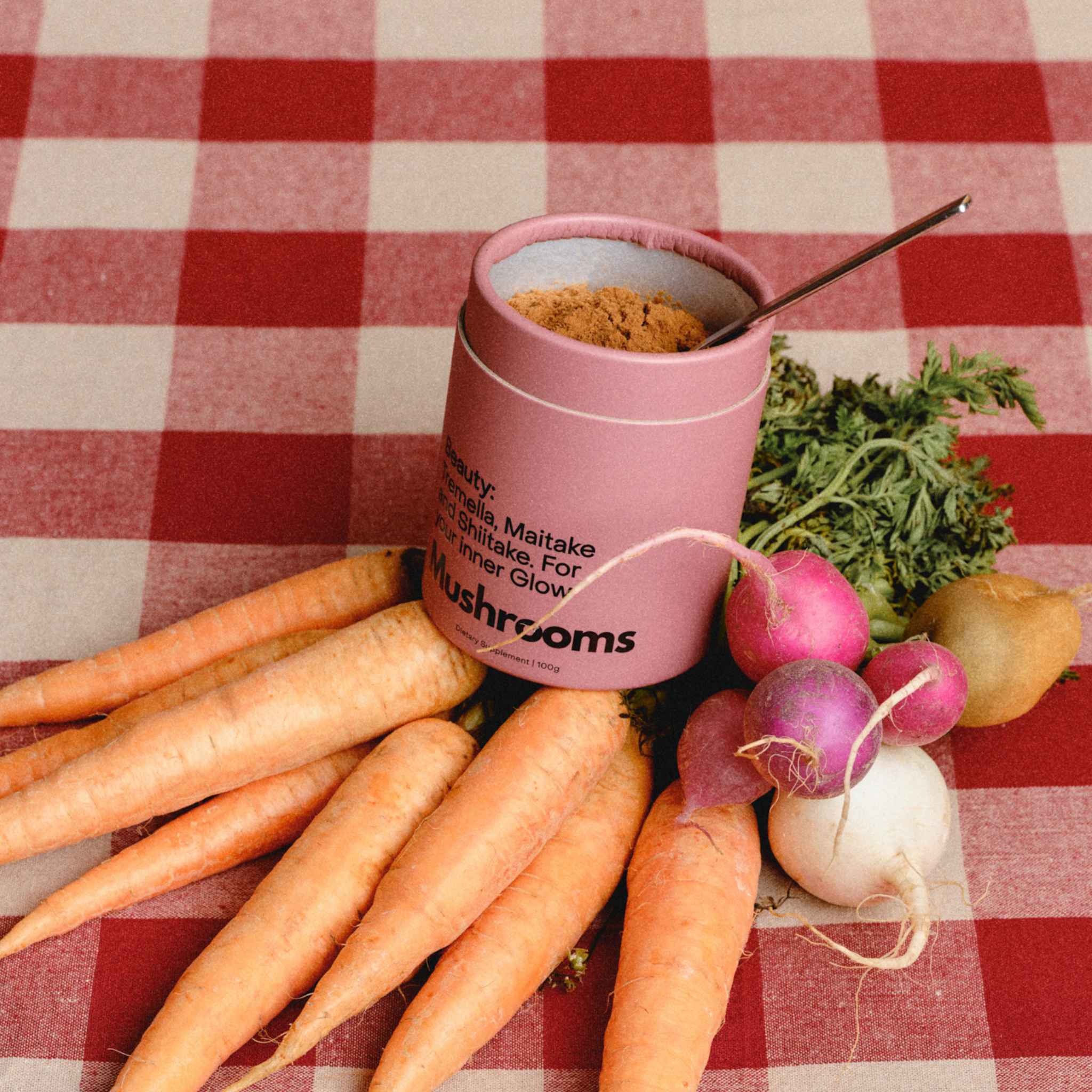 An open canister of the Mother Made Beauty Blend with a spoonful of the mushroom powder inside, styled on a red and white checkered tablecloth with carrots, radishes and a potato.