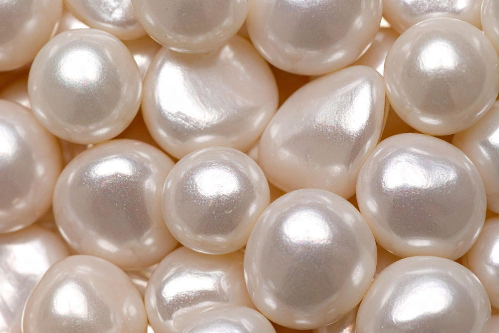 A close-up of glossy, irregular white pearls filling the frame use in natural beauty, health and wellness product when ground down into pearl powder, showing a soft pearlescent sheen and warm off-white tones.