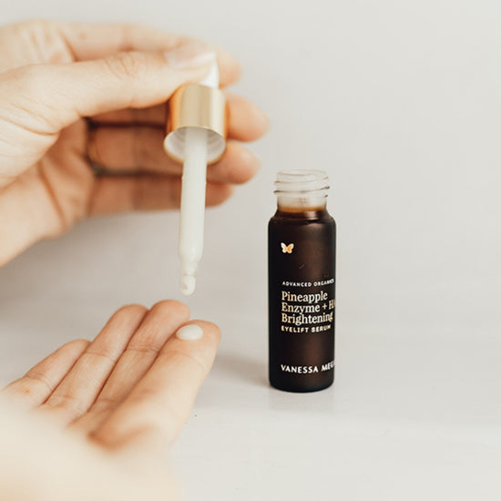 Vanessa Megan Pineapple Enzyme + HA Brightening Eyelift Serum with dropper dispensing serum onto hand, amber glass bottle in background, lightweight eye treatment for brightening, de puffing and hydration