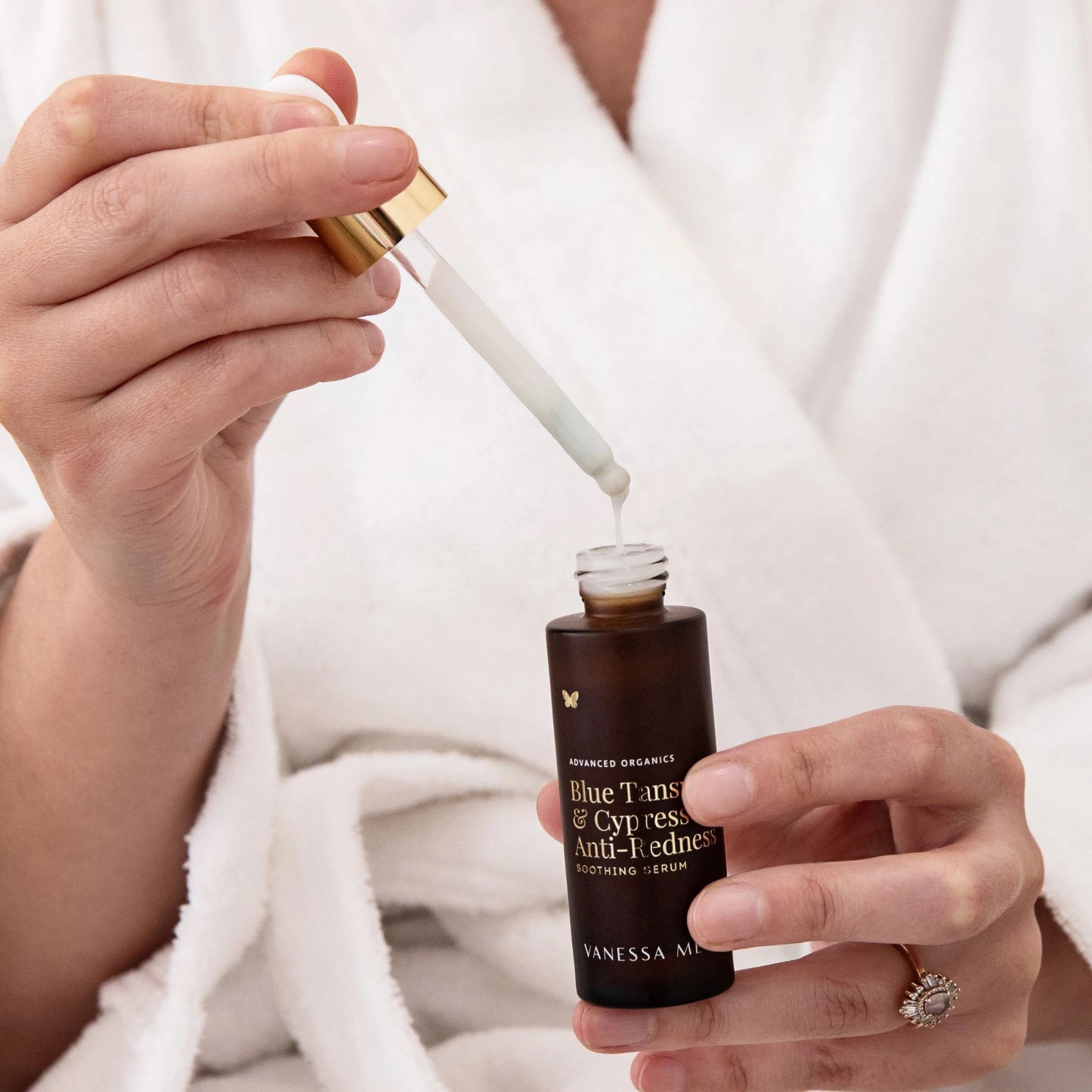 Hands holding of the formulation of the Vanessa Megan Blue Tansy and Cypress Anti-Redness Serum — lightweight, fast-absorbing milky serum dripping from dropper into amber glass bottle.
