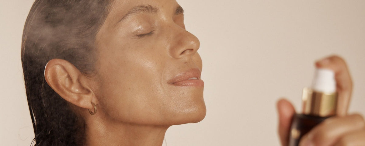 A close-up lifestyle shot of a woman with closed eyes, spraying a fine mist of the Vanessa Megan Rose Hydrosol Pore Refining Toner from a dark bottle with a gold collar and white pump her skin.