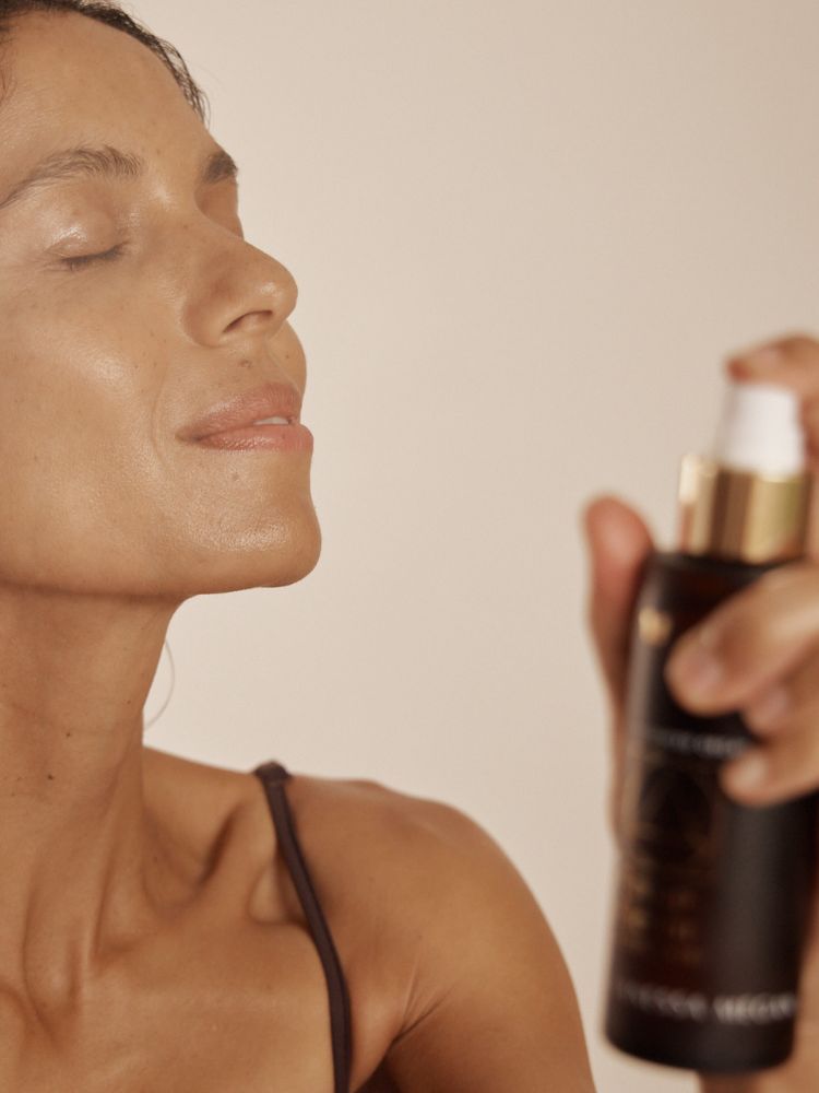 A close-up lifestyle shot of a woman with closed eyes, spraying a fine mist of the Vanessa Megan Rose Hydrosol Pore Refining Toner from a dark bottle with a gold collar and white pump her skin.