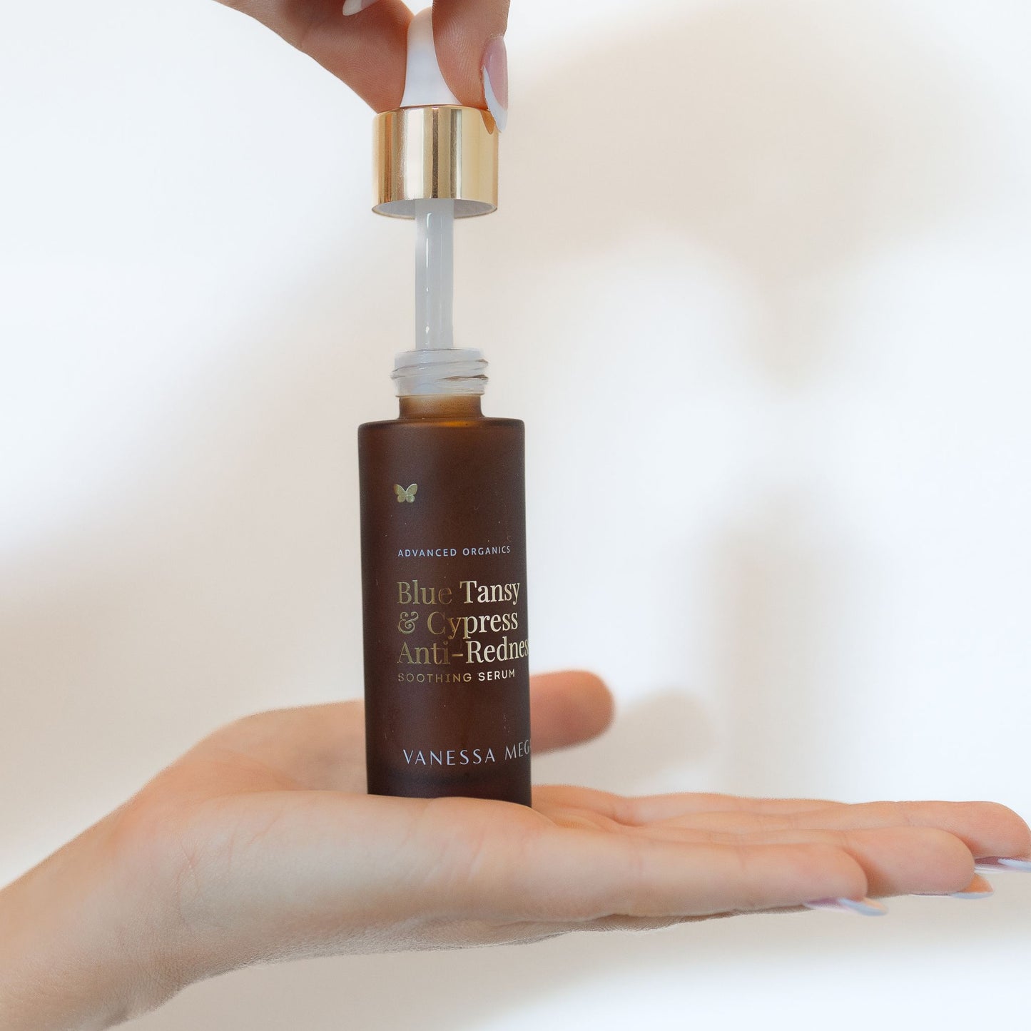 Vanessa Megan Blue Tansy and Cypress Anti Redness Serum bottle held in open hand with glass dropper lifted above, showing clear serum applicator against soft neutral background.