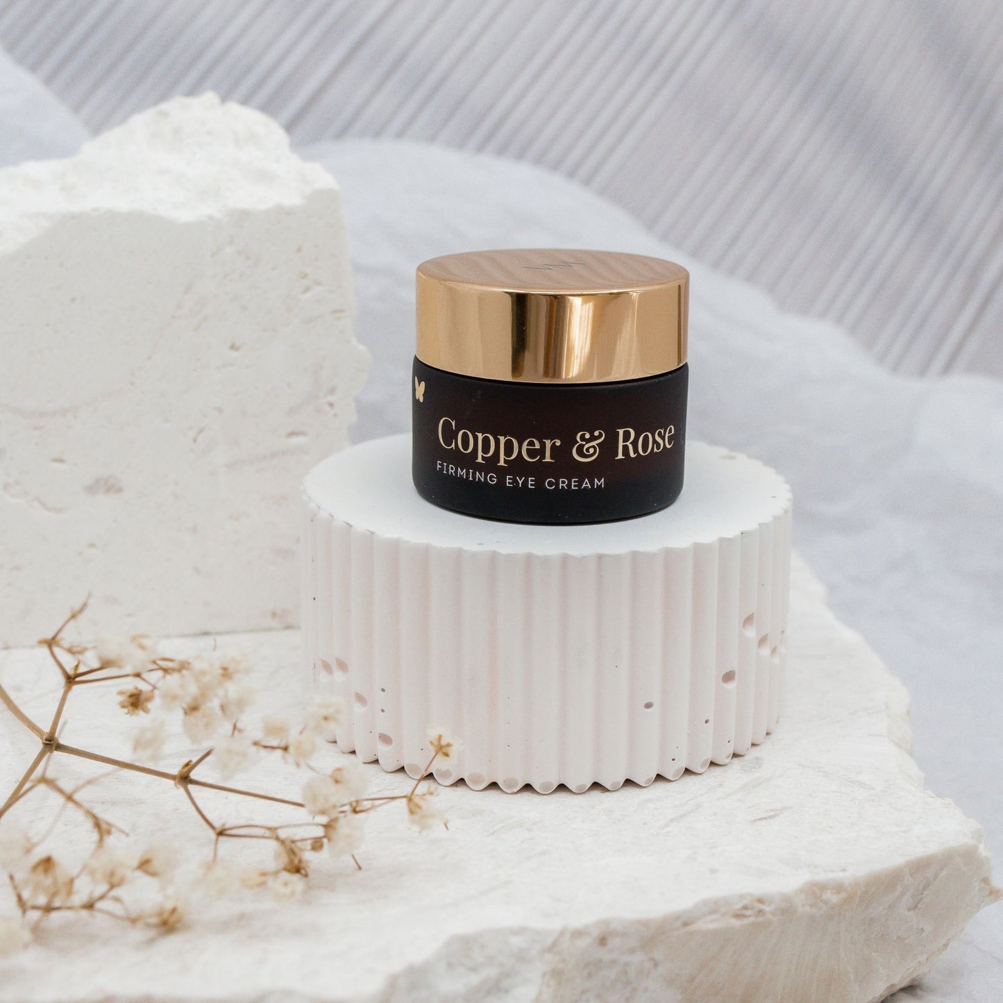 Vanessa Megan Copper and Rose Firming Eye Cream Jar styled on white pedestal with babies breath flower to the side.