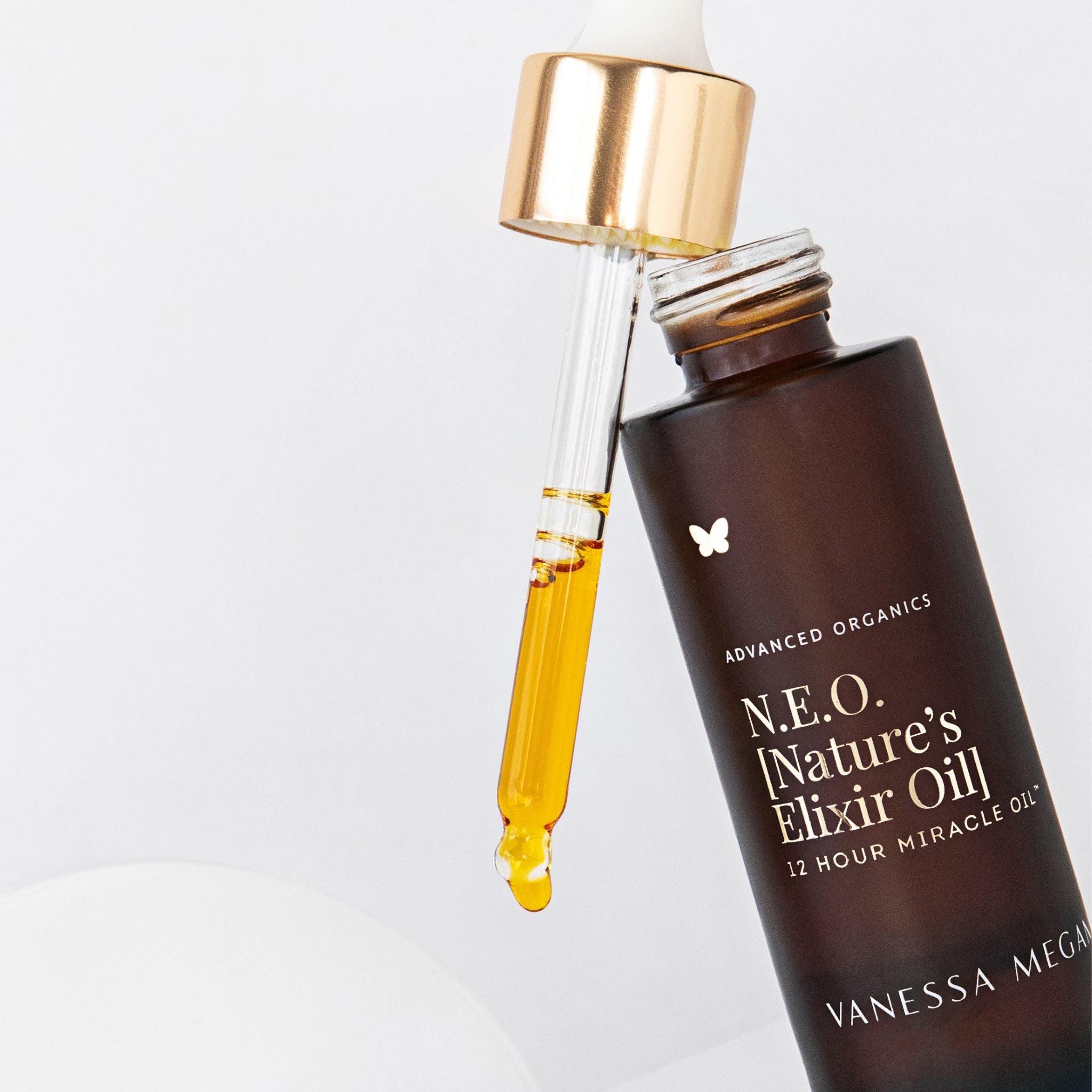 Vanessa Megan N.E.O Nature’s Elixir Oil 12 Hour Miracle Oil with glass dropper releasing golden botanical facial oil, close-up of amber bottle with gold cap on a clean white background.
