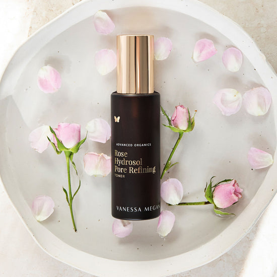 Vanessa Megan Rose Hydrosol Pore Refining Toner in amber glass bottle with gold cap, resting in a ceramic dish filled with water and scattered pink rose petals and fresh rose buds.