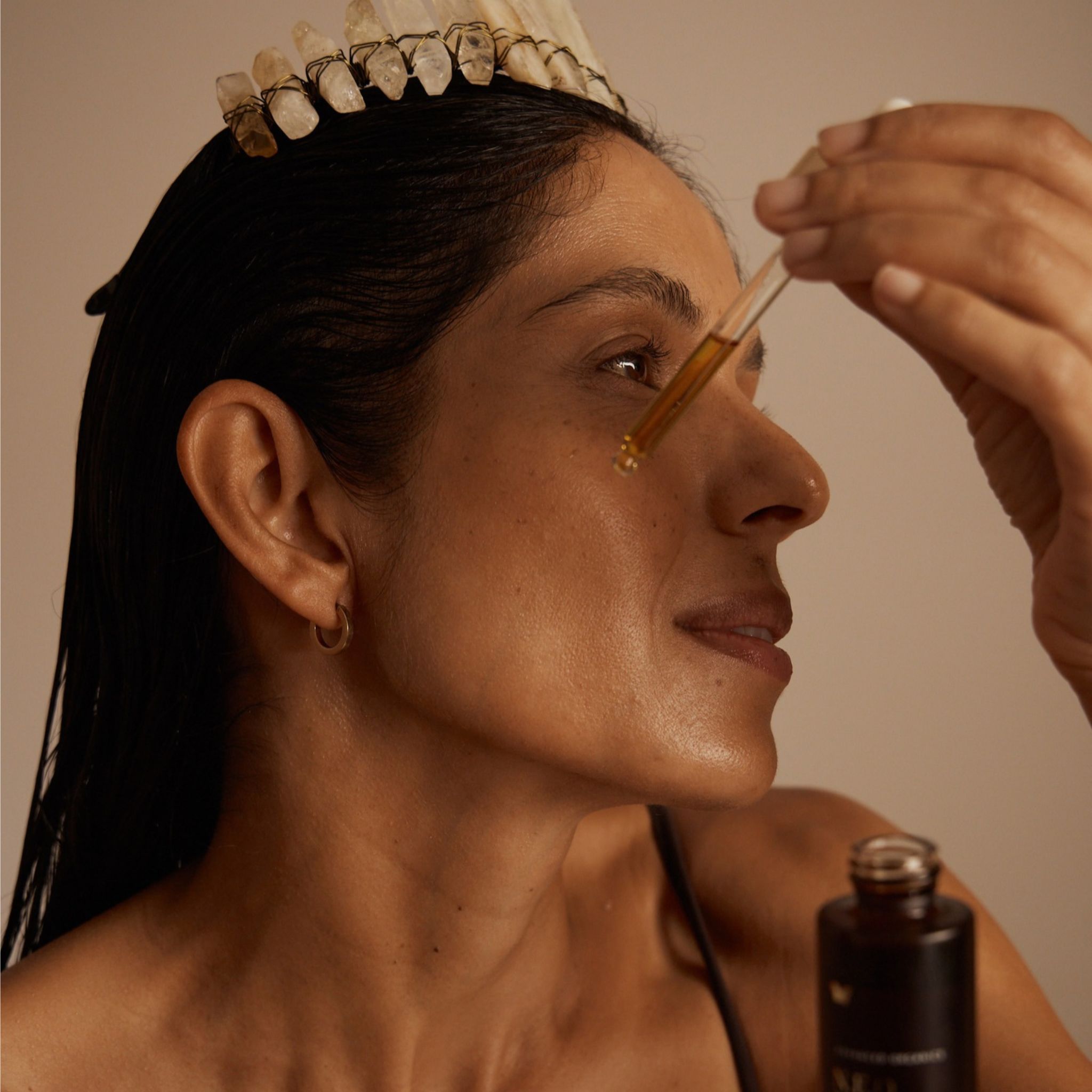 A woman with dark hair wearing a Crystal crown, applying a drop of Vanessa Megan N.E.O 12 Hour Miracle Oil
from a glass pipette held by a hand, with the bottle visible in the lower right corner against a warm beige background.