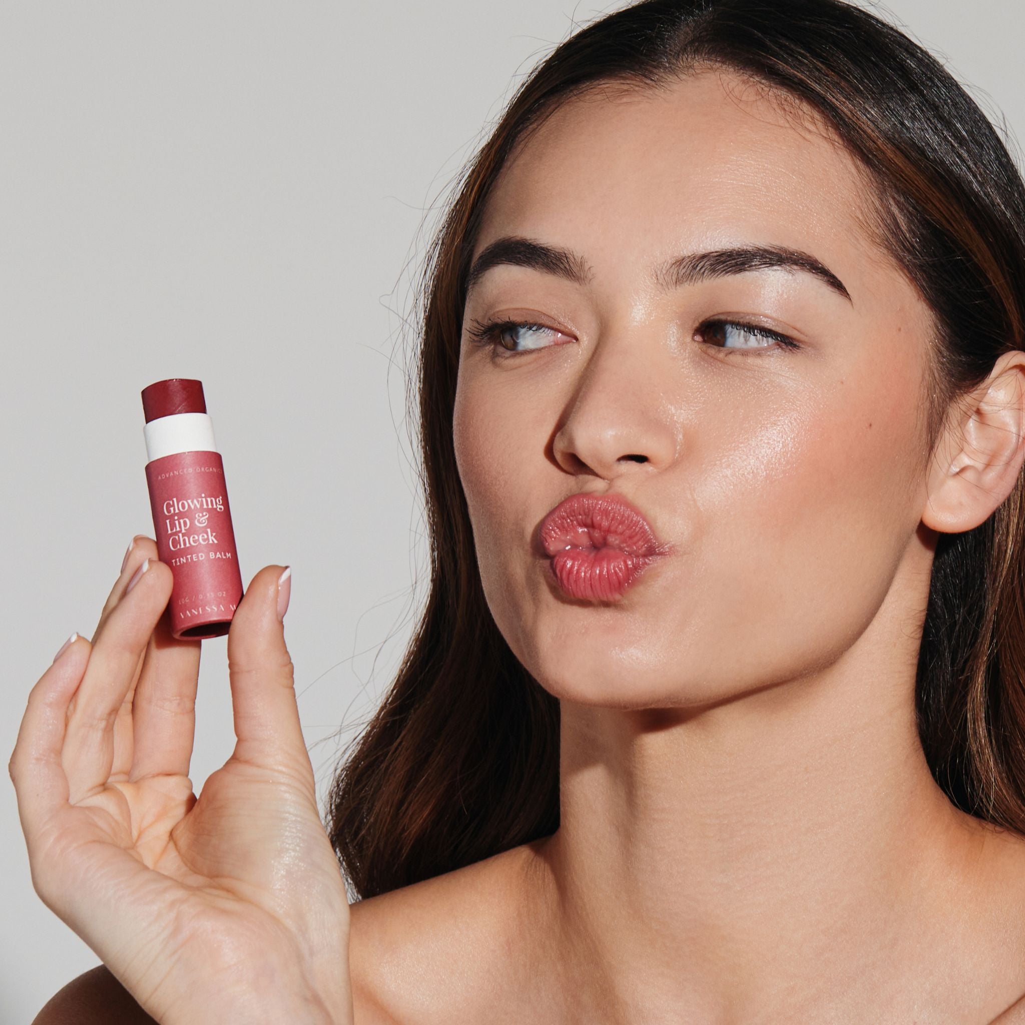Woman holding Vanessa Megan Glowing Lip and Cheek Tinted Balm close to face, blowing a kiss with soft rosy lips and natural luminous glow from clean organic makeup.