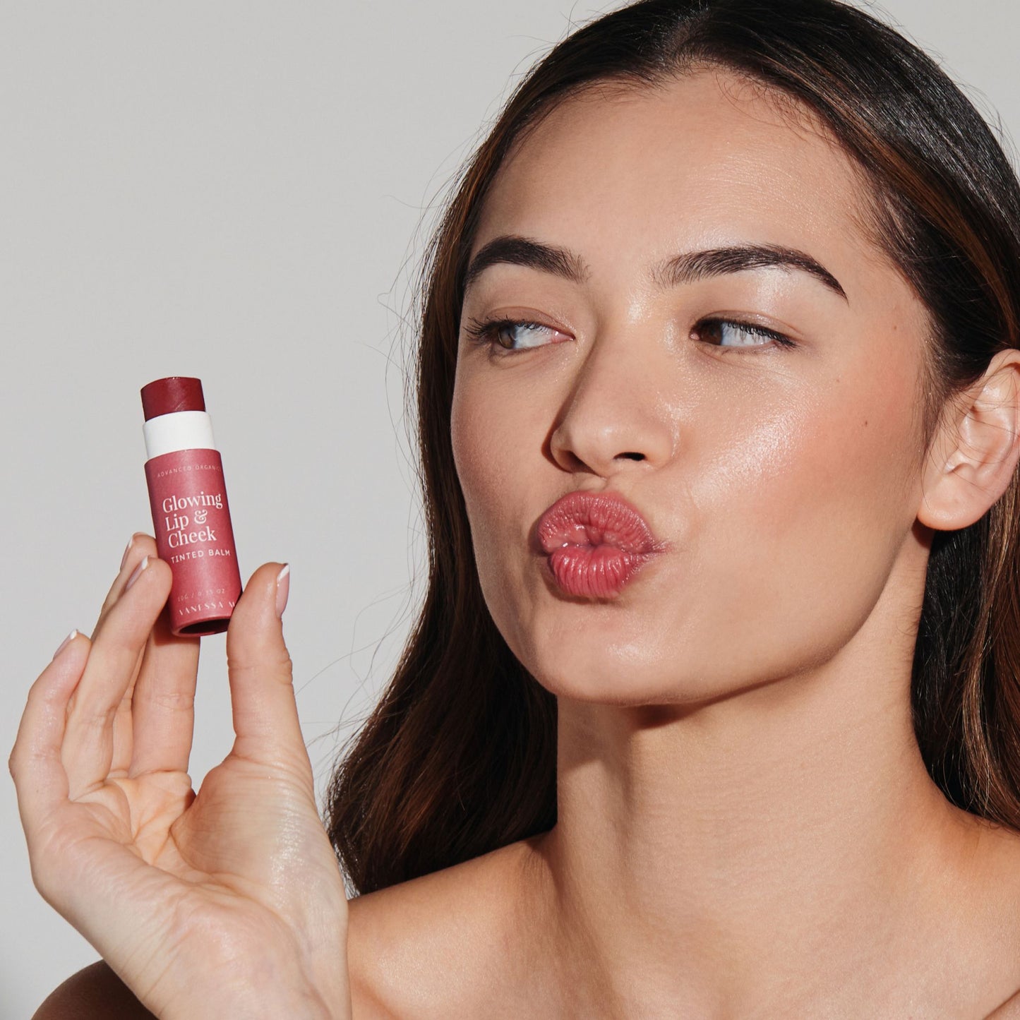 Woman holding Vanessa Megan Glowing Lip and Cheek Tinted Balm close to face, blowing a kiss with soft rosy lips and natural luminous glow from clean organic makeup.