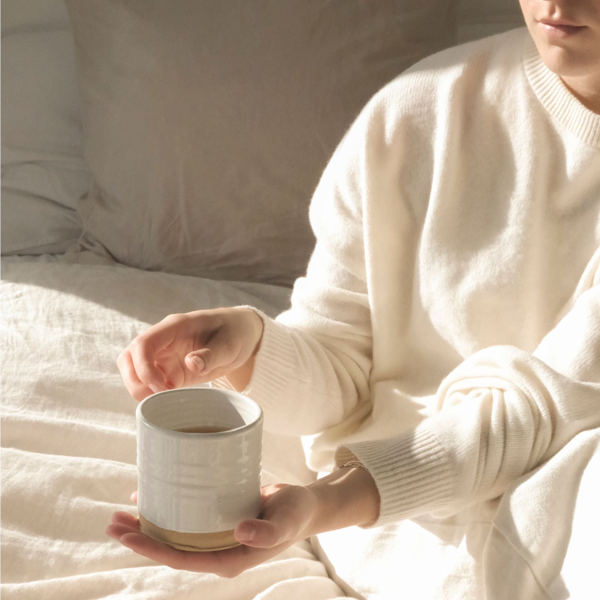 Woman holding a ceramic mug of On Sundays Earl Grey loose leaf tea in soft morning light on neutral linen bedding.
