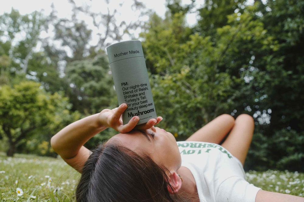 A woman lying on grass outdoors holds a Mother Made 220g card canister of Reishi, Shiitake and Turkey's Tail mushrooms against her face. Trees and grass are blurred in the background.